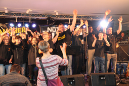 Gemeinsamer Auftritt der Internatskinder „Toni Orange and the Gang“ mit der Taucherband. Auf einer Bühne strecken viele Menschen Ihre Hände in die Höhe.